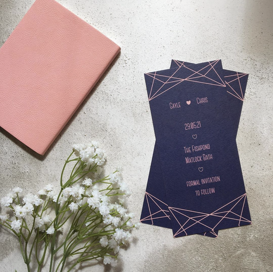 Blush and navy bookmark-style wedding invitation/save the date. The background colour is navy blue, and there is a geometric pattern on the top and bottom which is blush. The text is also blush.