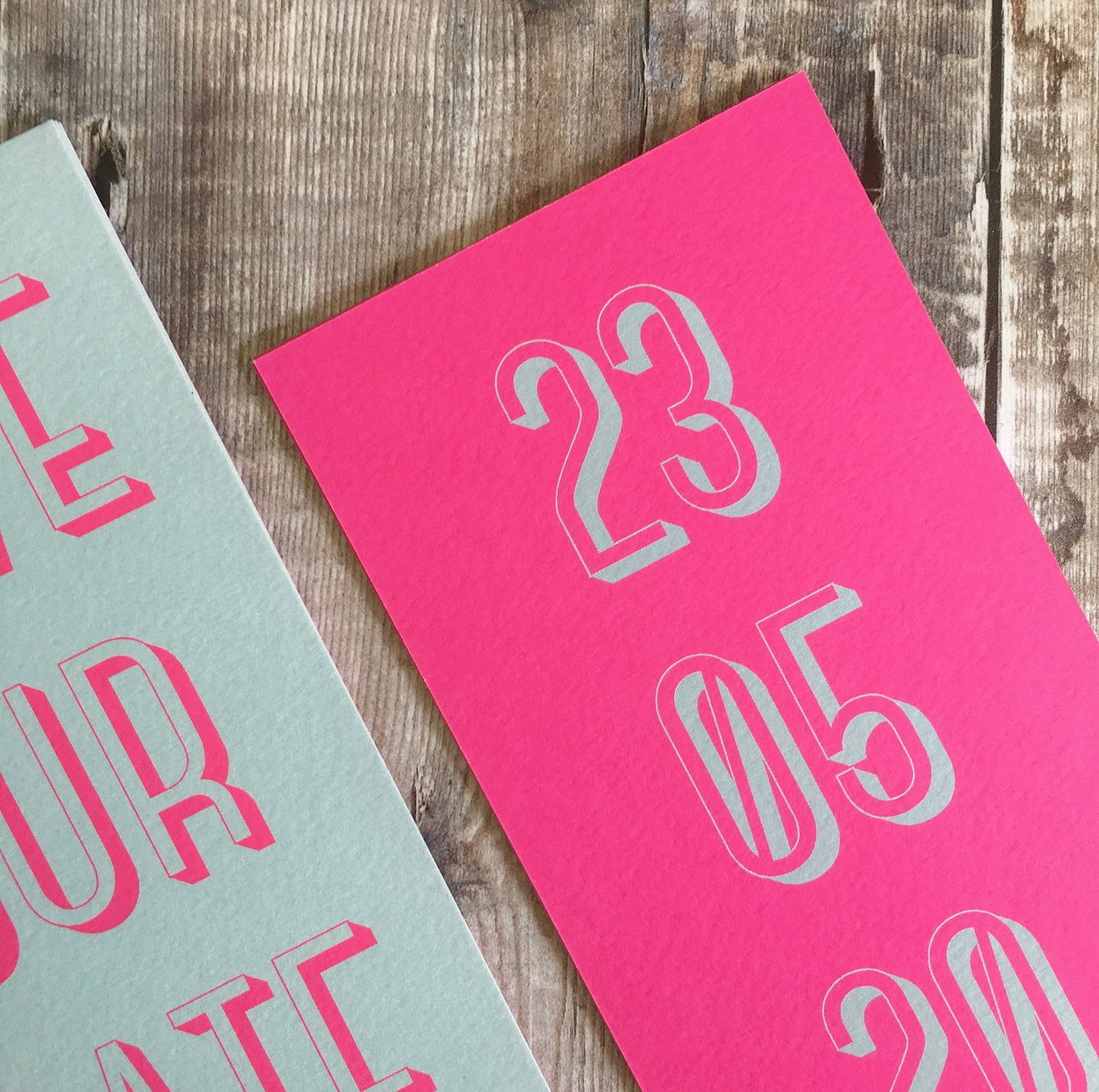 Close-up of the textured card. Bookmark-style wedding invitation/save the date. Back view. This has a pink fluorescent background, with the date of the wedding in a teal colour. The overall style is typographic and bold.