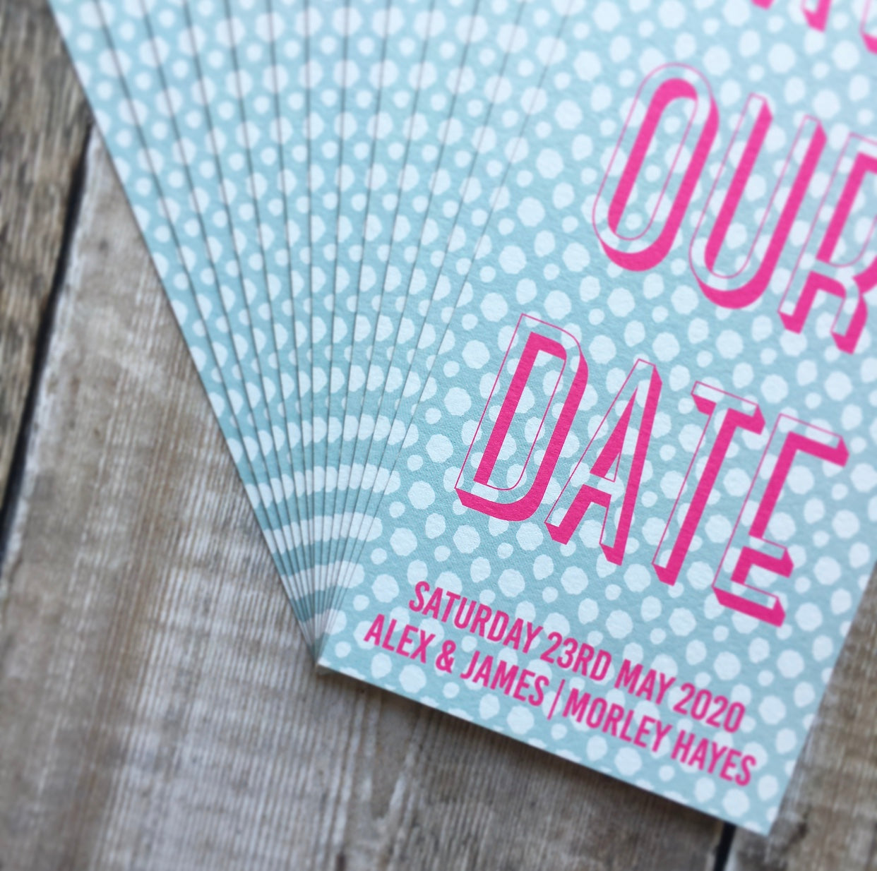 Close-up view. Bookmark-style wedding invitation/save the date. 99mm x 210mm. Pale blue background, with a white spotted pattern. Text is typographic, and written in fluorescent pink.